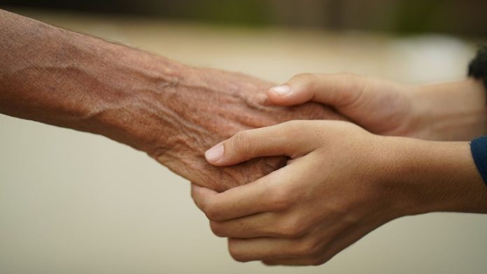 Na zdjęciu widać dłonie młodszej osoby i rękę starszej osoby. Młodsza osoba trzyma w swoich rękach dłoń starszej osoby.