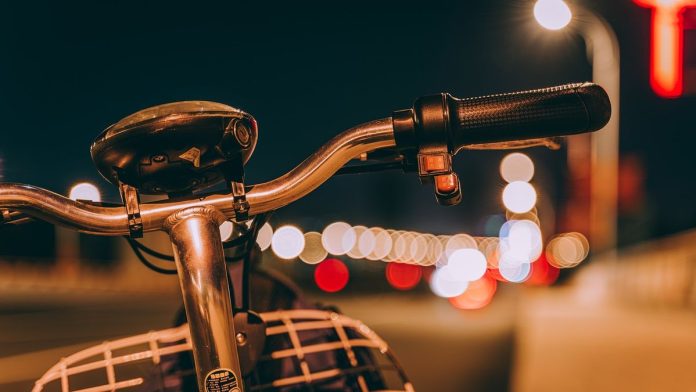 Zdjęcie przedstawia kierownicę roweru na tle świateł aut. Fotografię wykonano w nocy.
