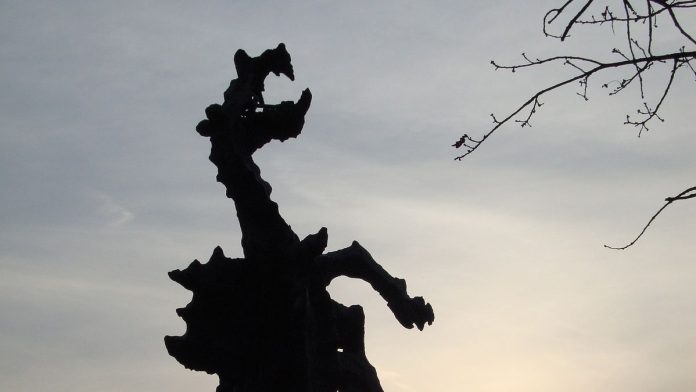 Zdjęcie przedstawia rzeźbę Smoka Wawelskiego na tle zachmurzonego nieba i gałęzi drzewa.